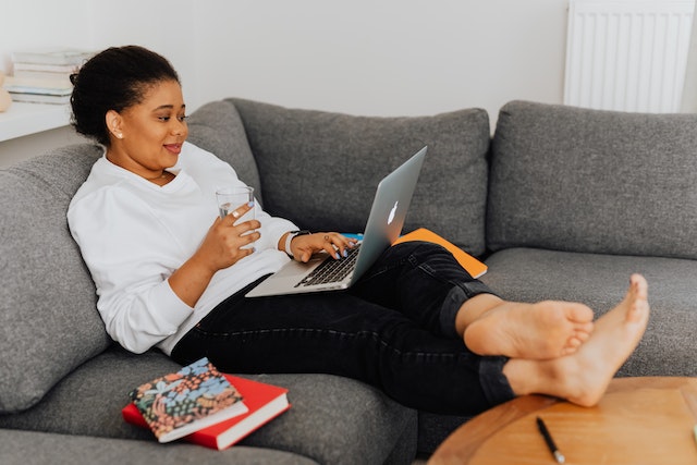 A woman sitting on her couch, leg stretched on the coffee table, a glass of water in hand with her laptop on her lap